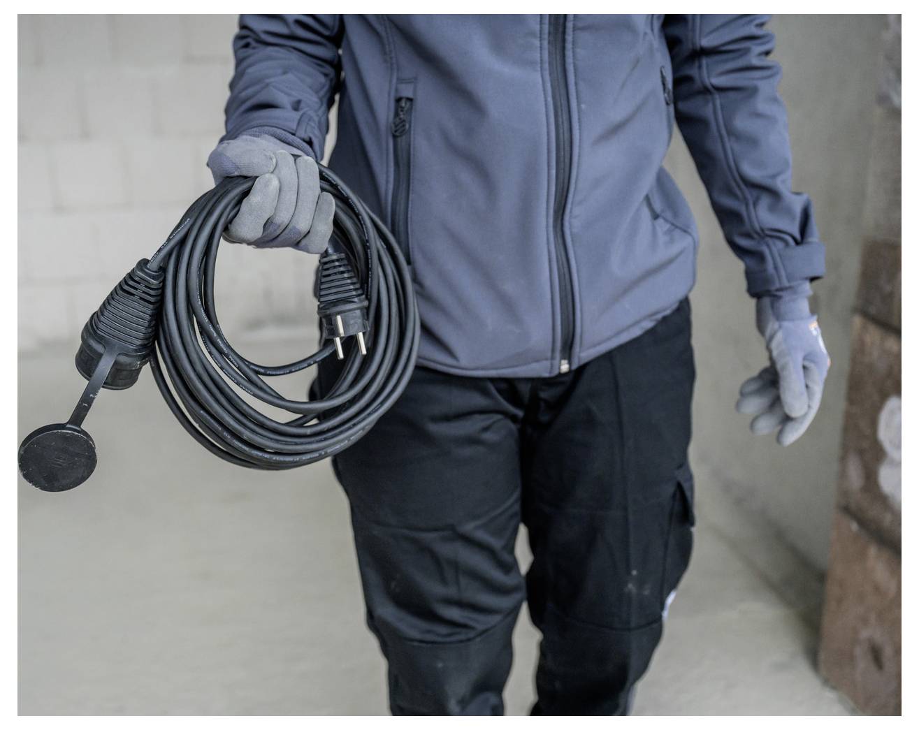 Person in work clothes holding a coiled electrical cord and plug, with a round black tool attachment dangling from it.