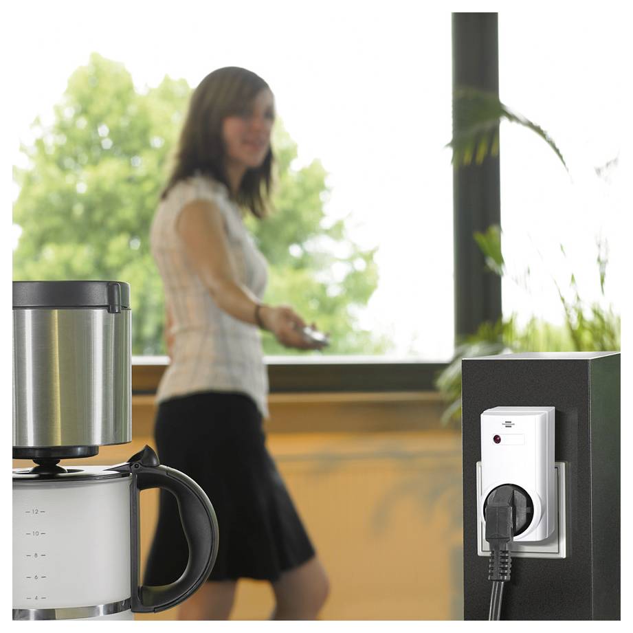 Office break room with a woman walking near a coffee maker and a plugged-in wall outlet in the foreground.