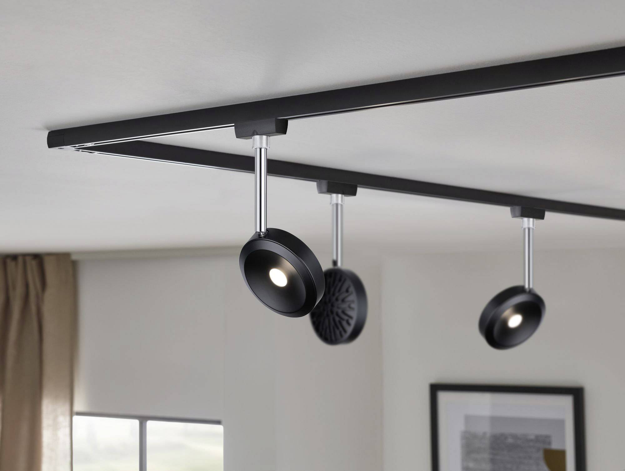 Three black ceiling spotlights on a track illuminate a room. In the background, a window with a curtain and a picture frame are visible.