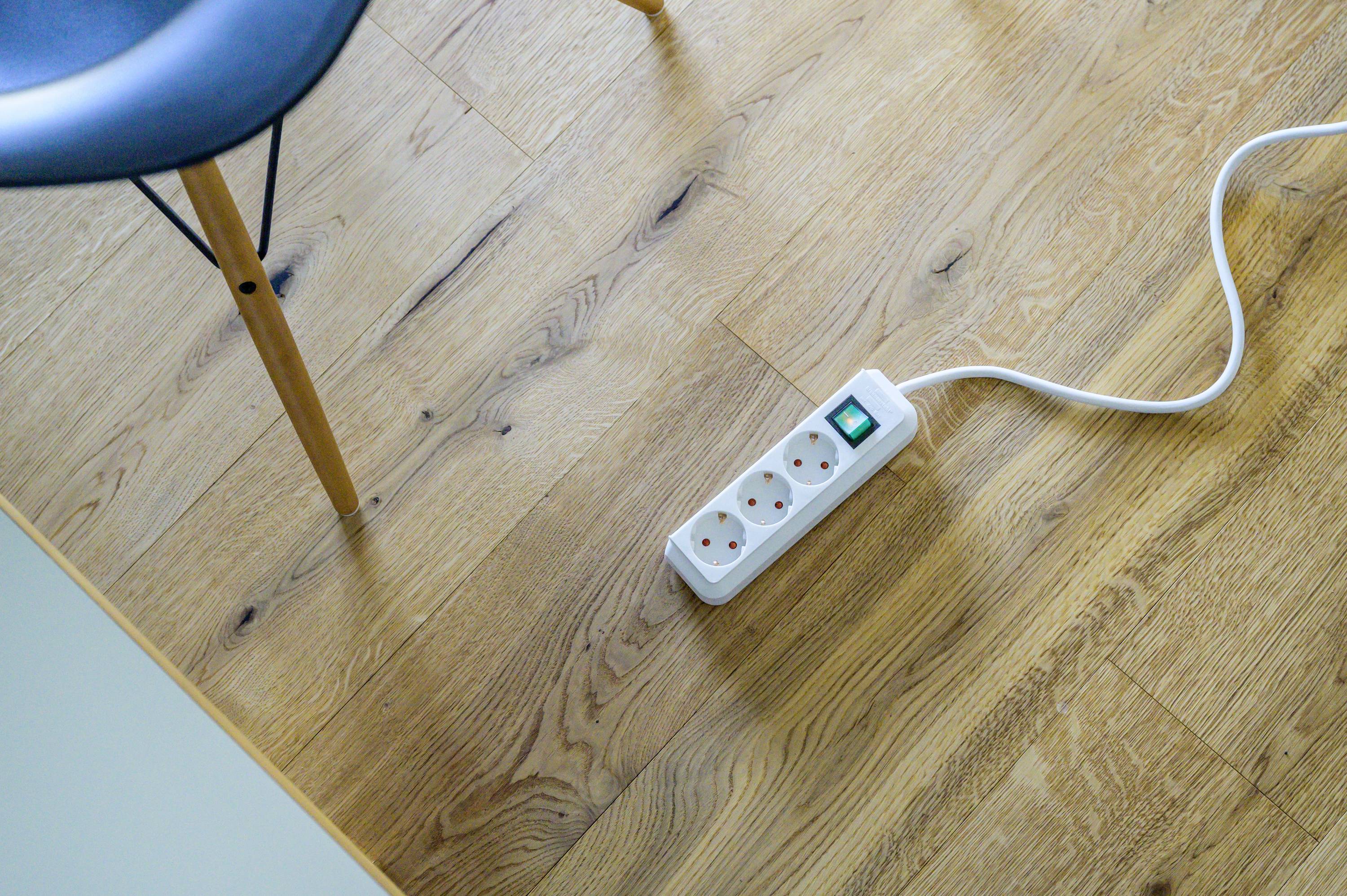 Multi-socket extension lead with an illuminated switch on a wooden floor next to a chair. One cable is plugged in.