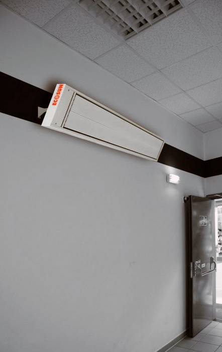 An air conditioning unit is mounted on the wall in a corridor. Next to it, an emergency exit door with an illuminated sign is visible.