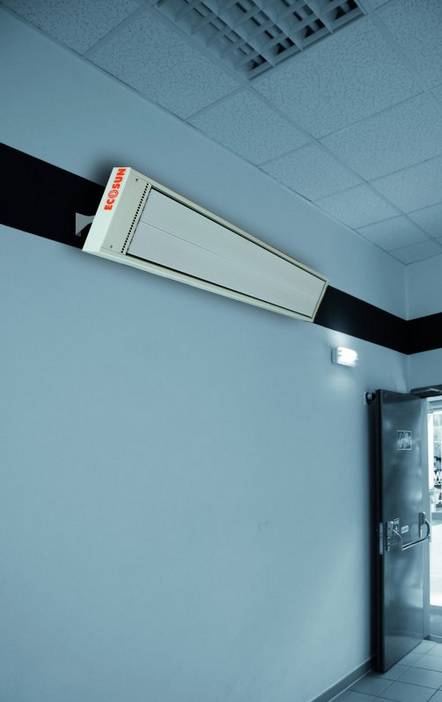 A heating radiator is mounted on the wall in a corridor. The door on the right in the image is slightly ajar, and there is an emergency light.