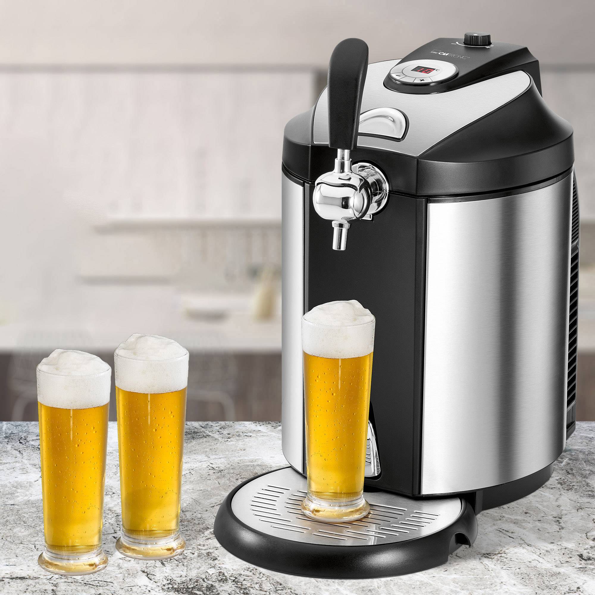 A silver beer tap stands on a table. Three glasses filled with light beer and foam are next to it. A blurry kitchen is in the background.