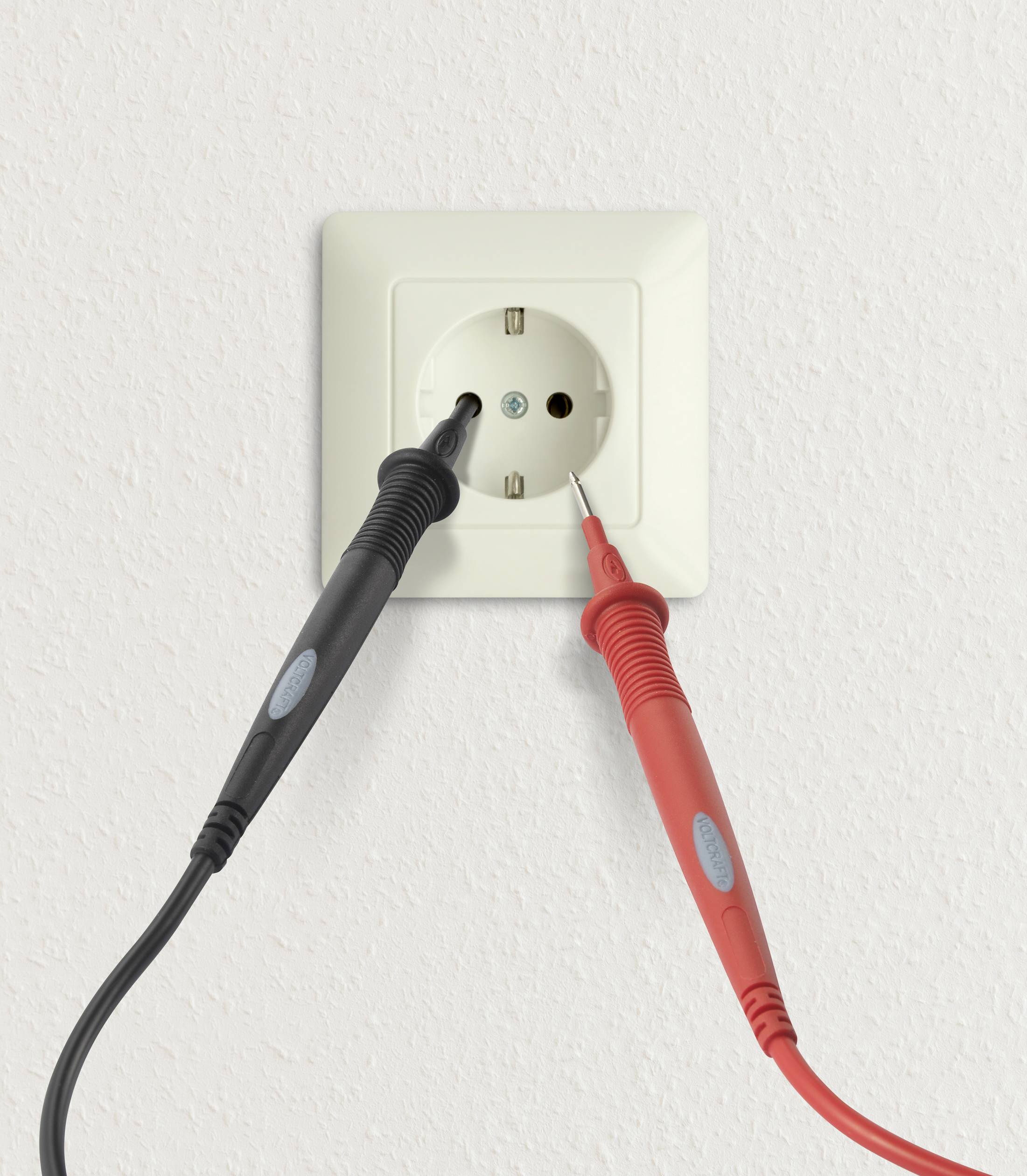 A current measuring device with red and black cables measures voltage at a white socket in the wall.