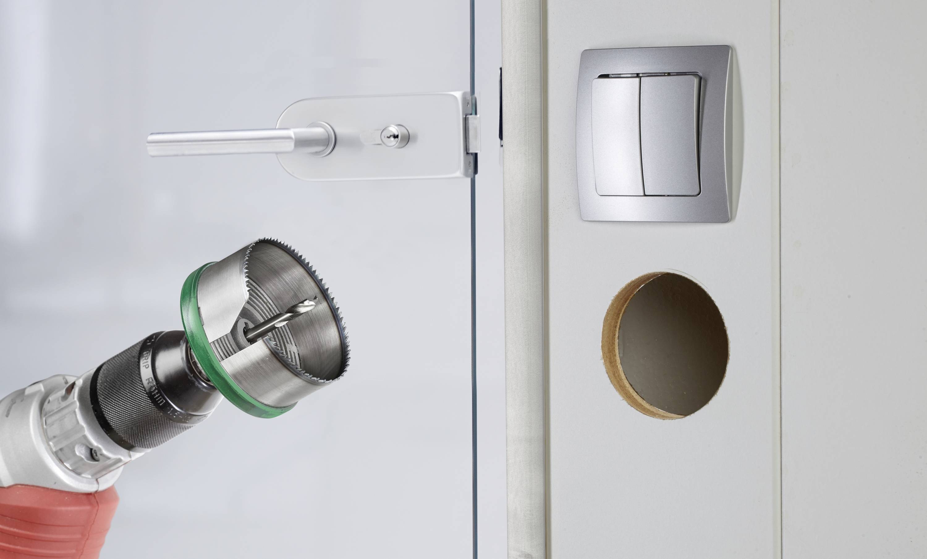 Drill with hole saw attachment near a glass door with a metal lock; a light switch and an opening in the wall beside the door frame.