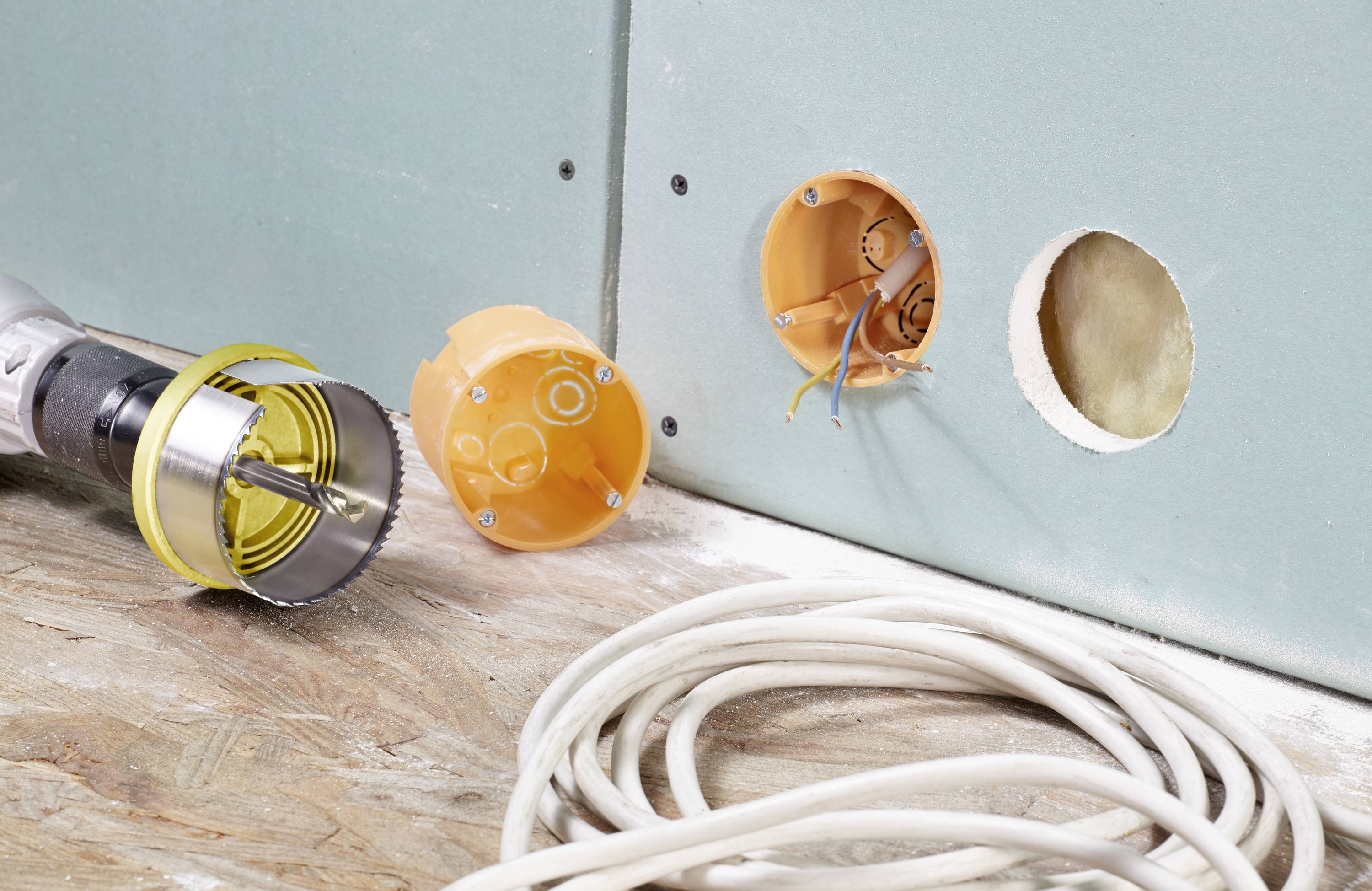 Drill with hole saw and electrical back boxes on a plasterboard wall; electrical cables on the floor. Construction site scene.