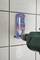 A drill is drilling a hole in a tiled wall, using a blue and red drill template holder as a guide.