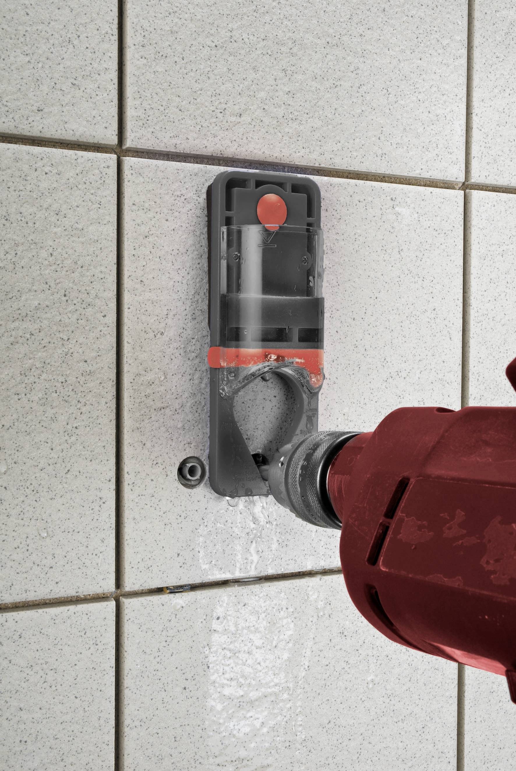 A red drill is boring into a wall with light-coloured tiles, using a grey dust extraction device.
