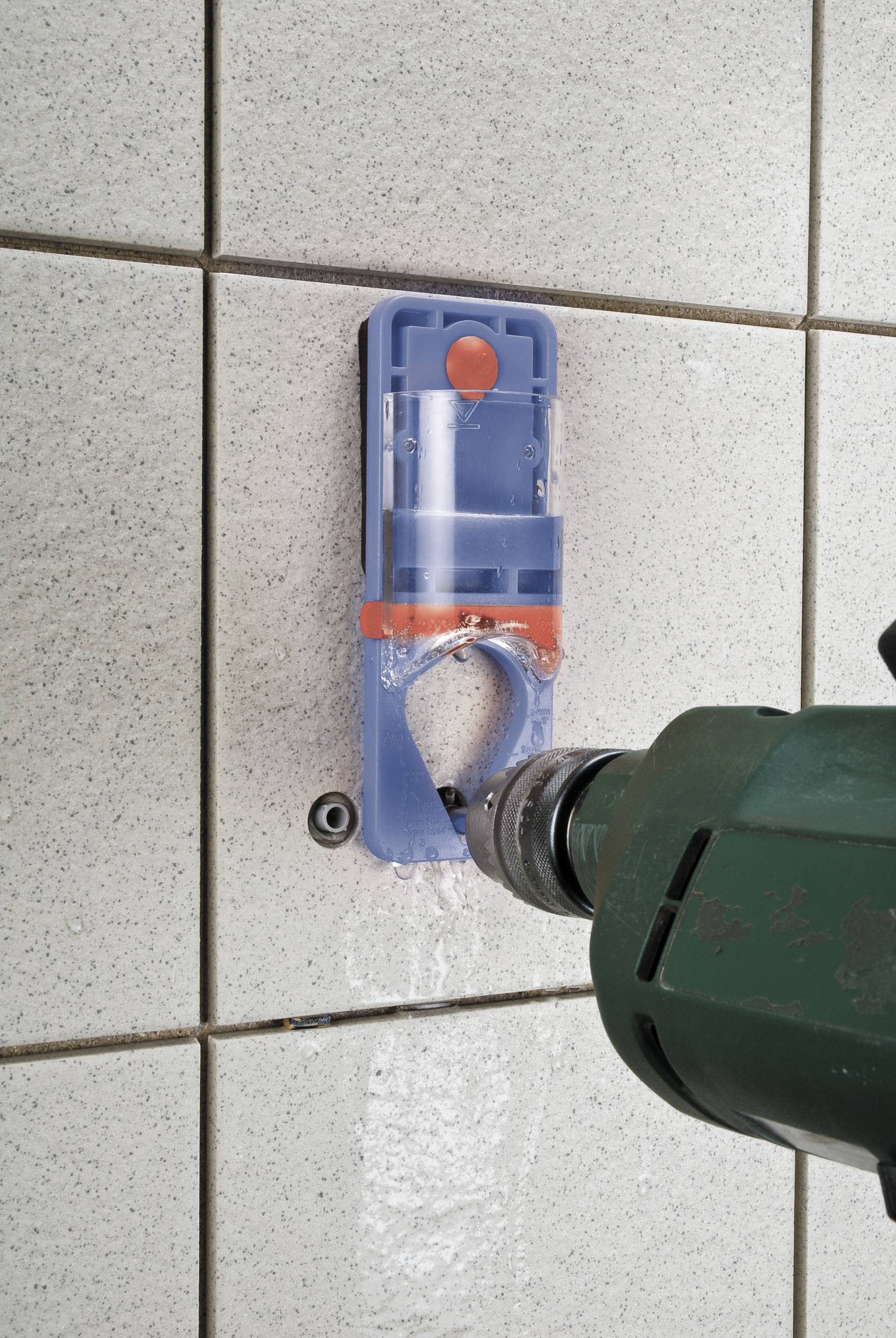 A green drill penetrates a tiled wall with a blue guide template supporting the drilling process.