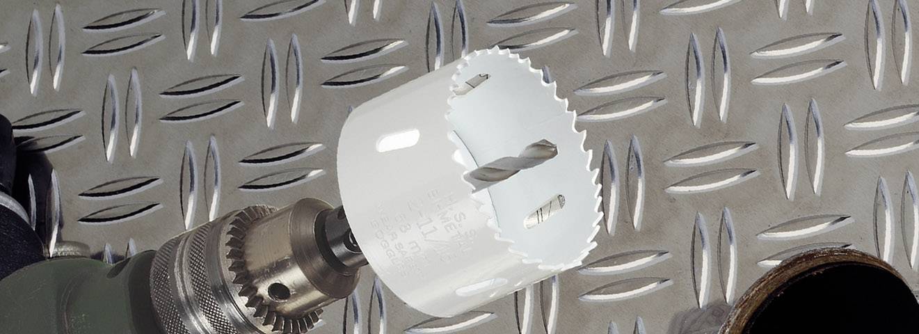 A drill with a hole saw attachment lies on a grooved metal surface. Part of a drill machine is visible on the left.