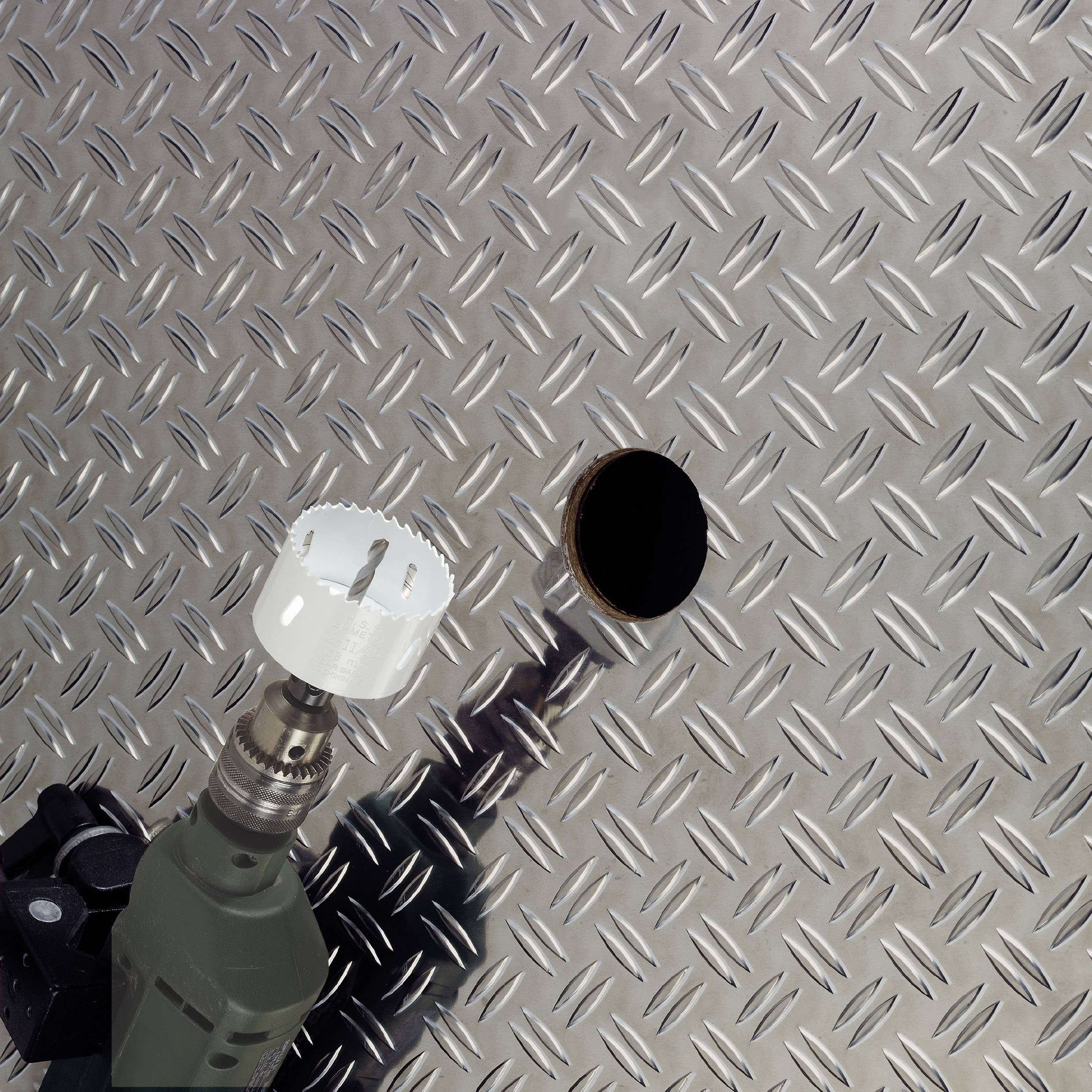 Drill with hole saw attachment next to a circular hole in a corrugated metal sheet, demonstrating metalworking.