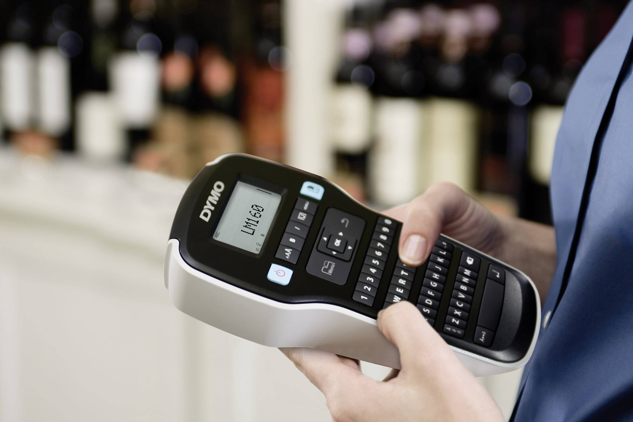 A person is holding a labelling device with the word 'LIVING' printed on it. Wine bottles are blurred in the background.