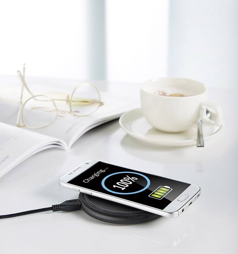 A smartphone is resting on a wireless charger with a 100% charged battery. Next to it are a coffee cup, a pair of glasses and a book.