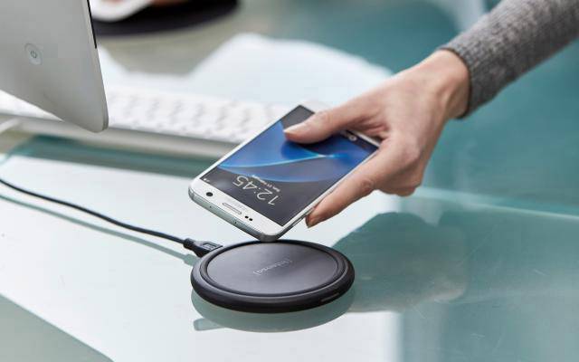 A hand holds a smartphone over a wireless charger on a desk. The display shows '12:45'.