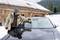 A man is scraping ice off a car windscreen in front of a wooden cabin in the snow. He is wearing a thick jacket and hat to protect himself from the cold.
