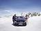 Two people are standing in front of a Mercedes in a snow-covered mountain landscape. One person is wearing a purple jacket, the other is wearing striped clothing.