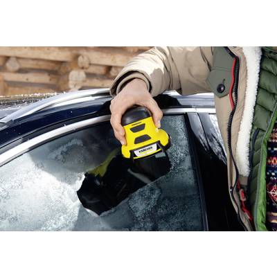 A person is removing ice from a car windscreen using an electric ice scraper. The car is black and there is a wooden wall in the background.