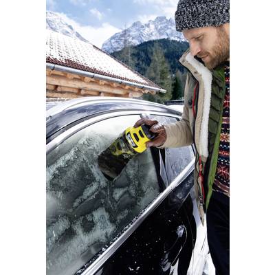 A man removes ice from a car windscreen using an electric scraper in wintry conditions. Mountains are visible in the background.