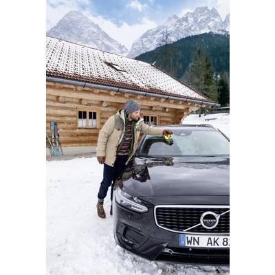 A man is scraping ice off the windscreen of a grey car in the snow in front of a wooden cabin with mountains in the background. Skis are leaning against the cabin.