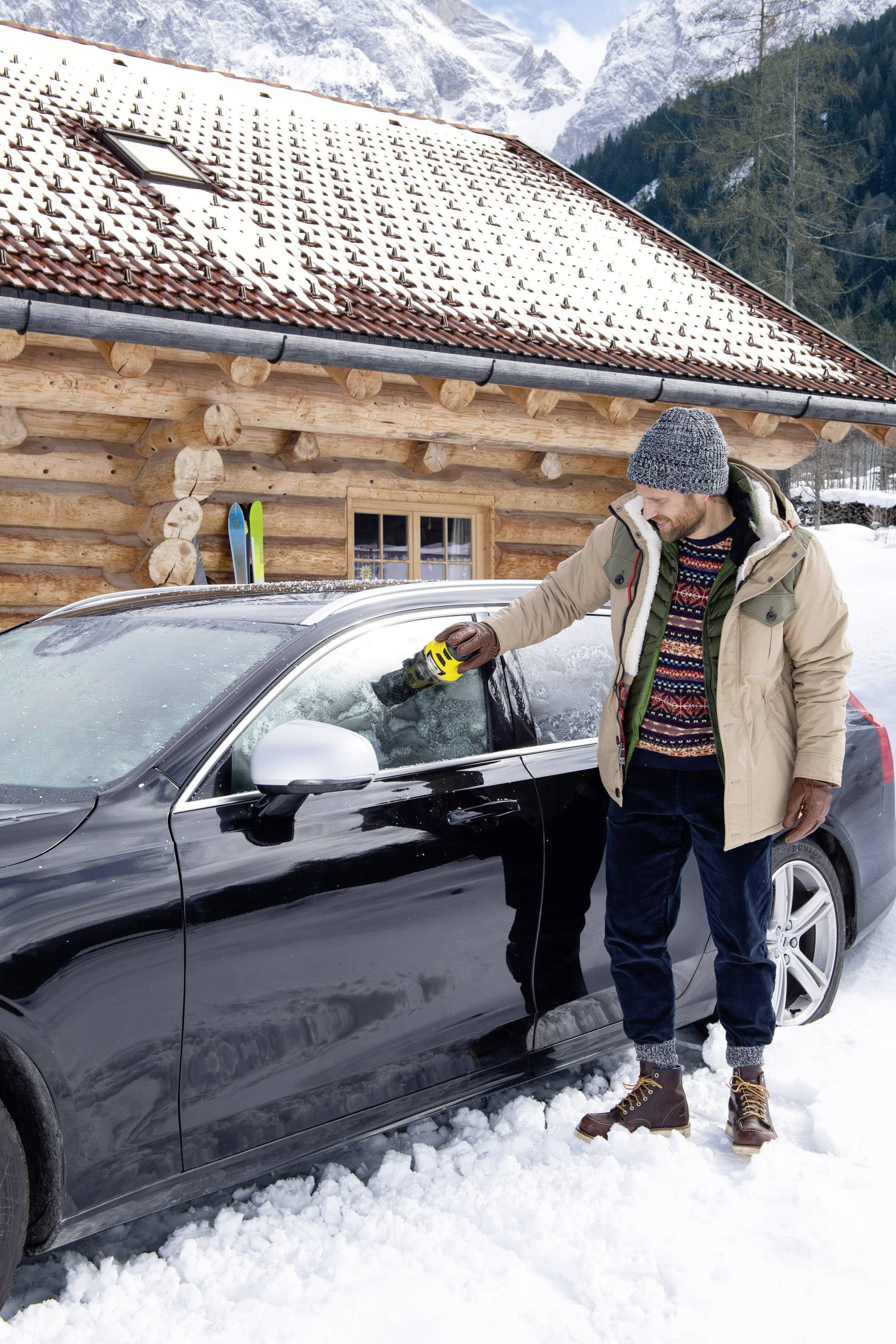 A man stands in the snow beside a car, removing ice from the windscreen with an ice scraper, with a wooden house in the background.