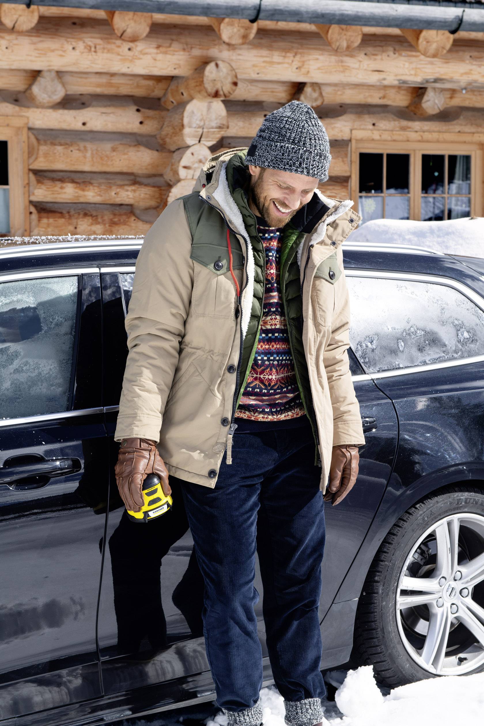 A man is smiling beside a car in the snow, warmly dressed in a woolly hat and jacket, holding a yellow device in his hand. A wooden cabin is in the background.