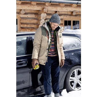 A man is smiling beside a car in the snow, warmly dressed in a woolly hat and jacket, holding a yellow device in his hand. A wooden cabin is in the background.