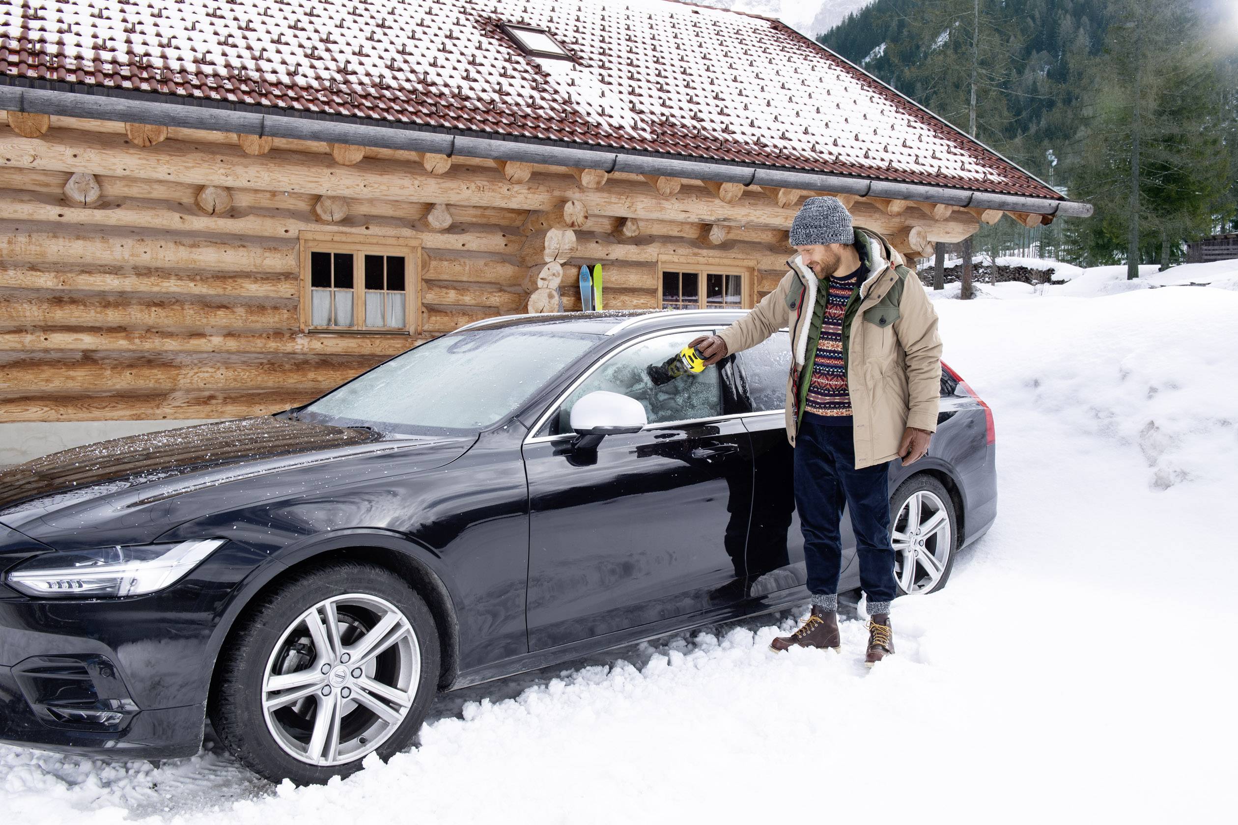 A man in a winter coat stands in front of a snow-covered car, removing snow from the window. A wooden cabin is visible in the background.