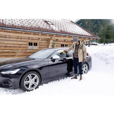 A man in a winter coat stands in front of a snow-covered car, removing snow from the window. A wooden cabin is visible in the background.