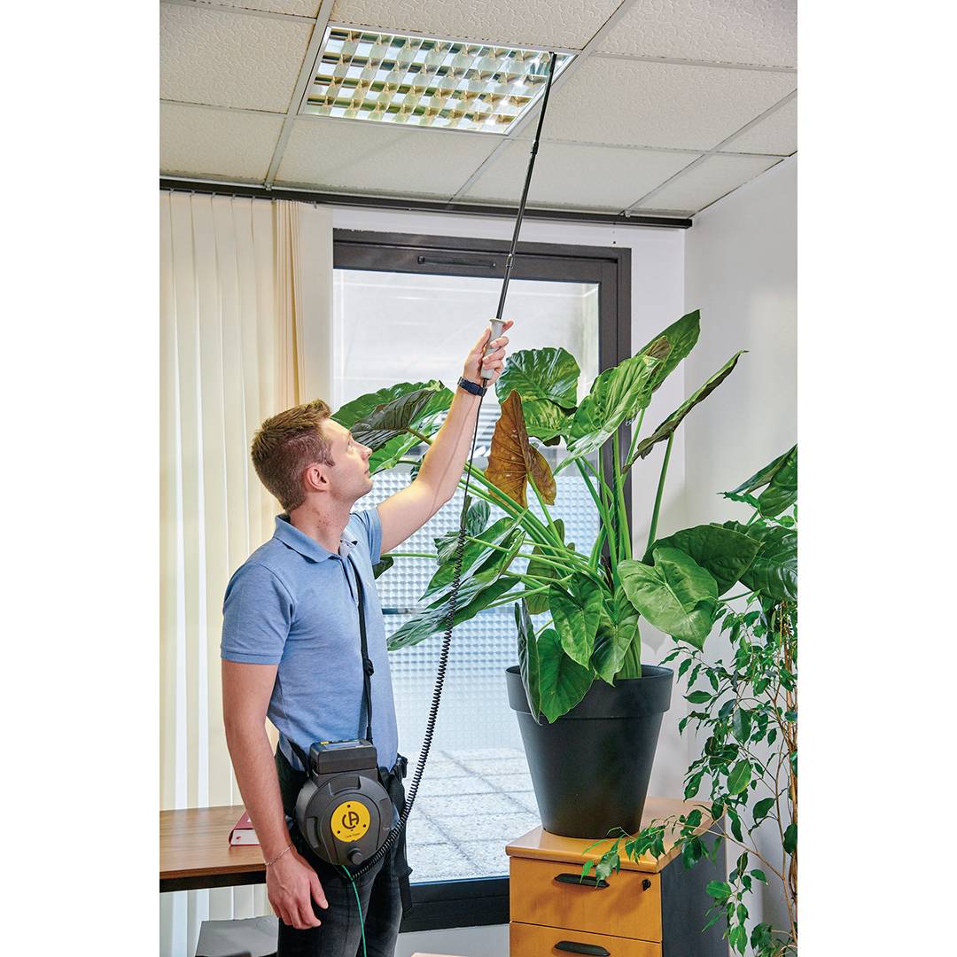 Man in blue shirt changing ceiling light with long tool in office. Large plant in foreground.