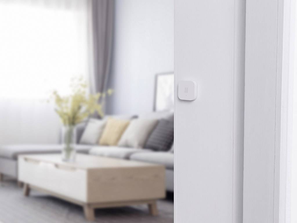 A modern living room with a sofa, decorative cushions and a coffee table. A square sensor is mounted on the wall.