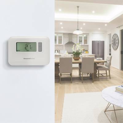 A modern living room with a table and chairs. On the wall, a thermostat with a digital display showing '20 degrees'. Kitchen in the background.