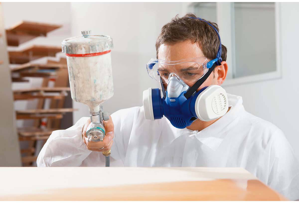 A man is wearing protective clothing and a respiratory mask while using a spray gun to paint a surface in a room.