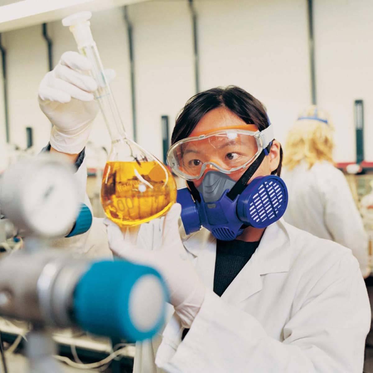 A scientist in the laboratory is wearing safety goggles and a respiratory mask, holding a swirling flask containing an orange liquid.