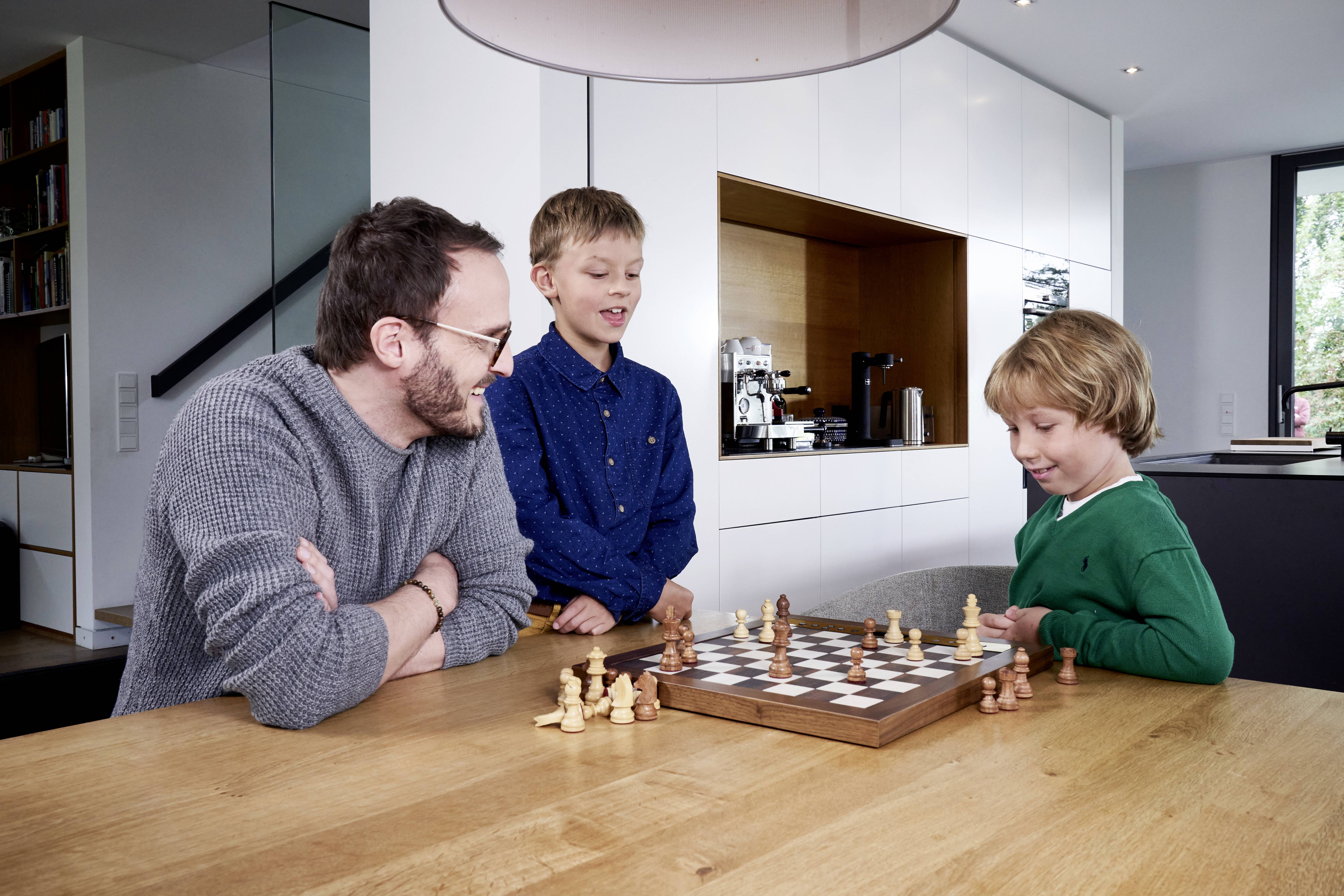 An adult and two children are playing chess at a wooden table in a modern room. All are looking intently at the board.