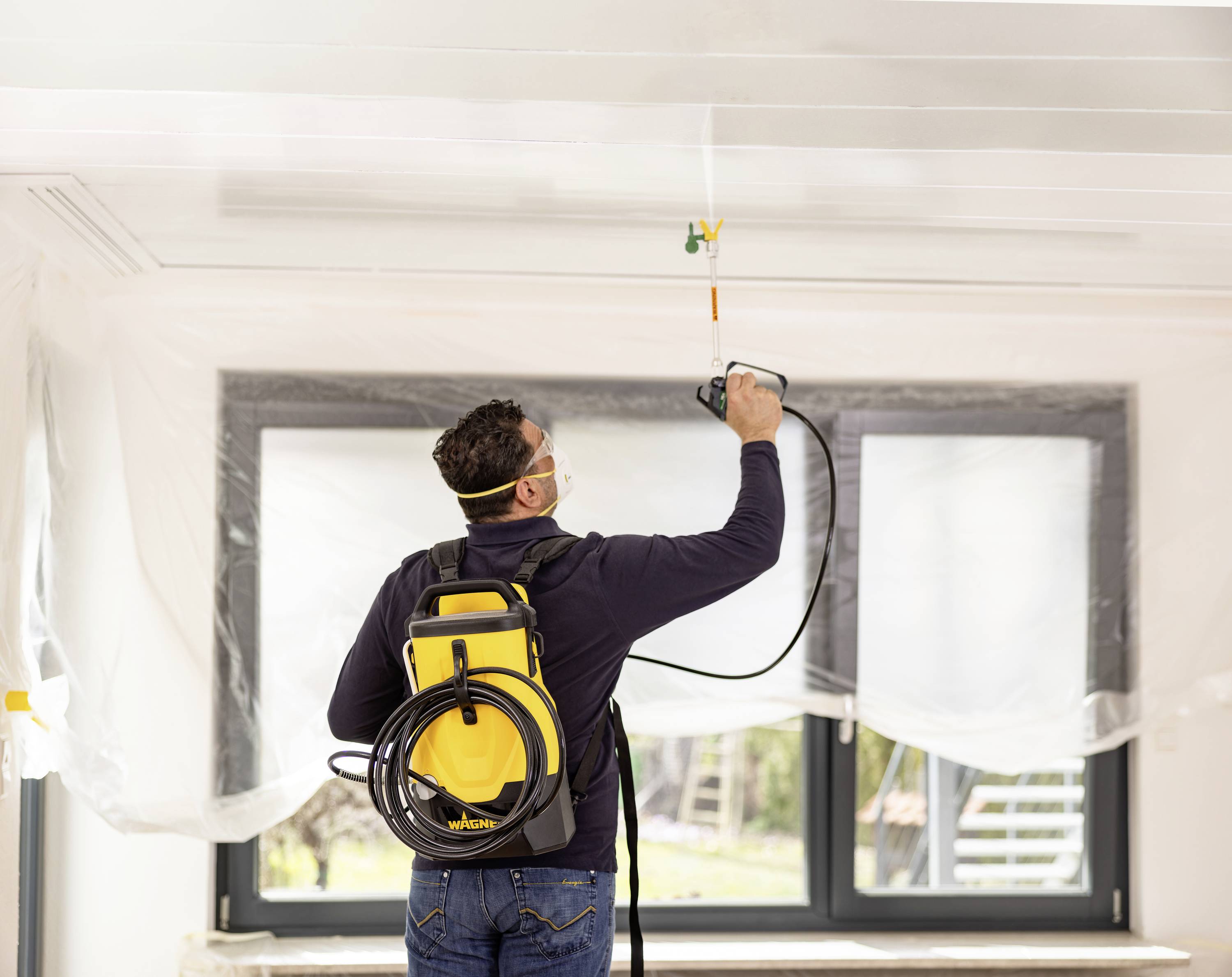 A man is spraying the ceiling of a room with a paint sprayer. He is wearing safety equipment, including safety goggles and a respirator.