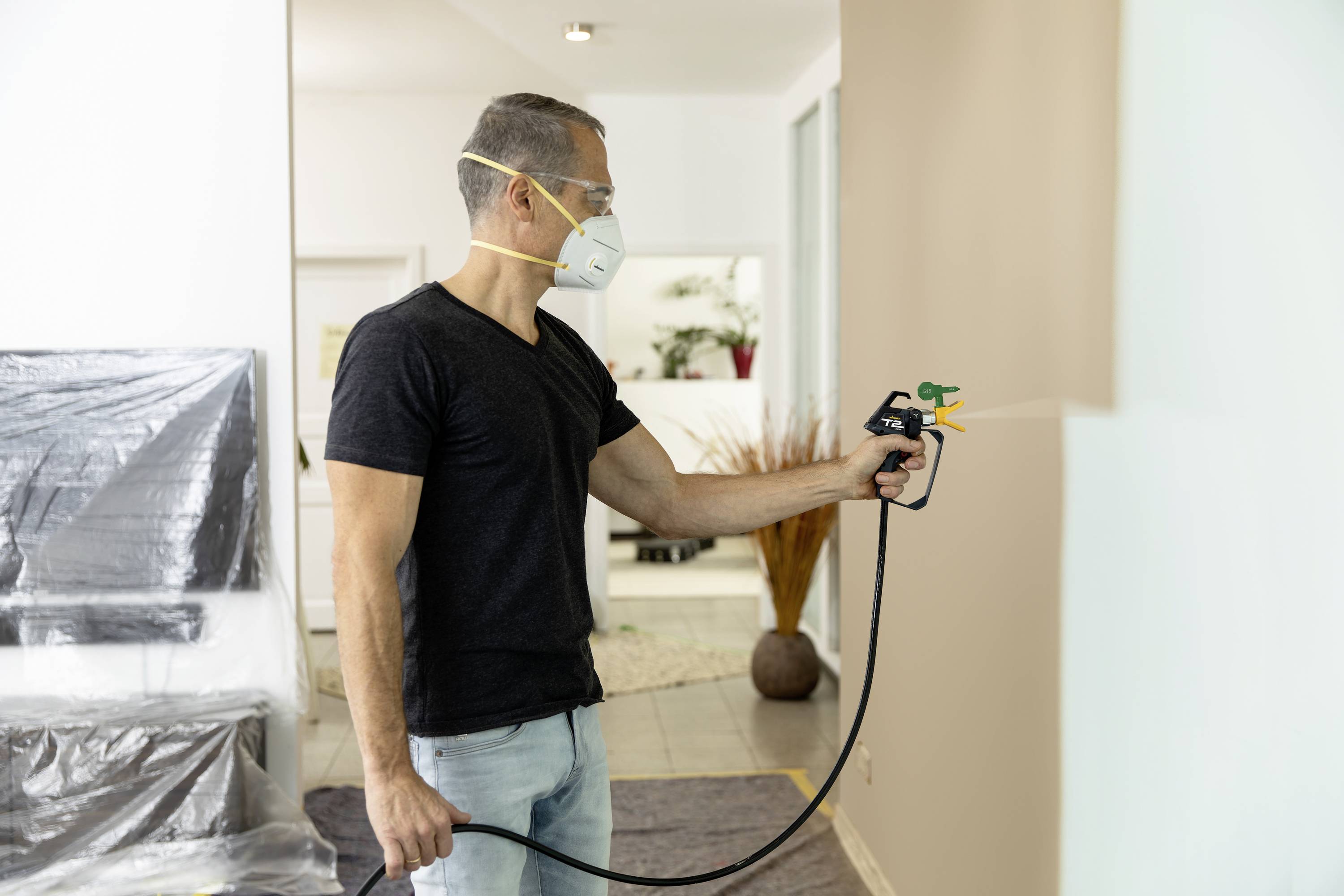 A person wearing a protective mask is spraying paint with a spray gun onto a wall in a modernly furnished room.