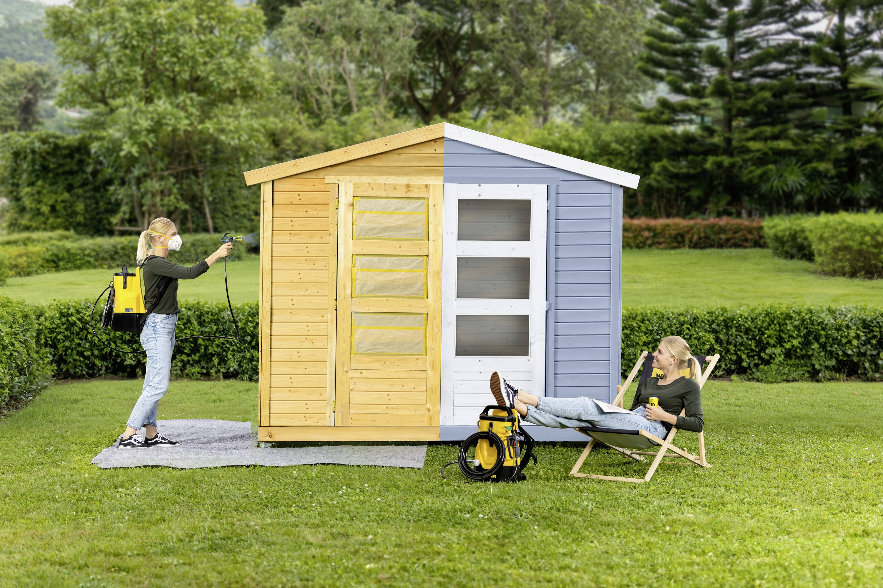 A woman is spraying a small, two-coloured garden shed yellow, while another relaxes in a deck chair. Grass and trees in the background.