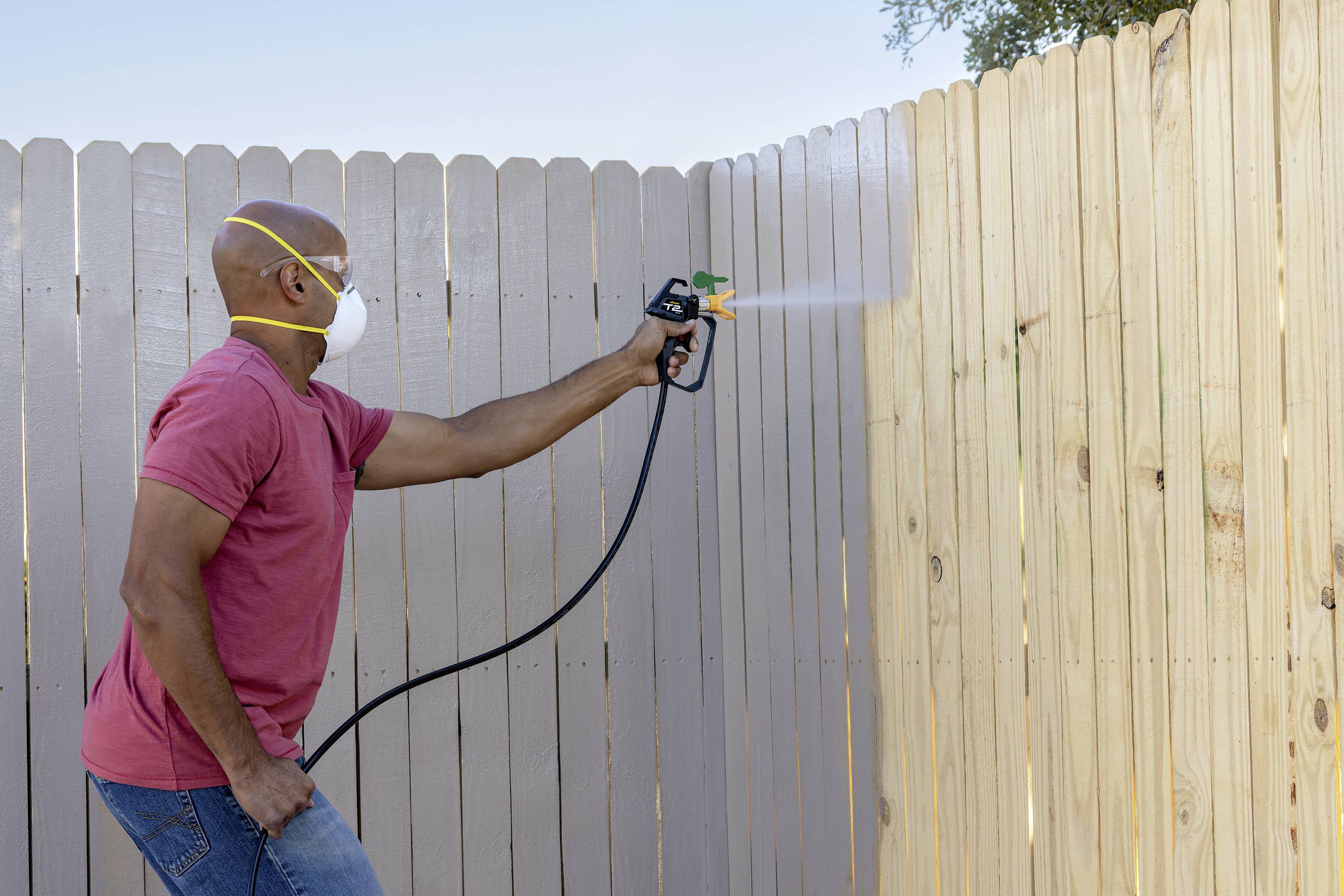 A person is spraying paint onto an outdoor wooden fence using a paint spray gun. They are wearing a protective mask.