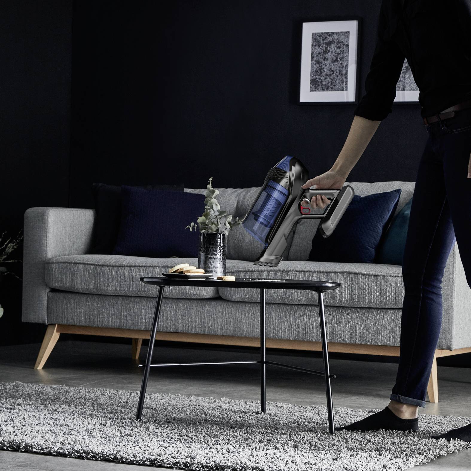 A person is hoovering the carpet in a living room with a cordless vacuum cleaner, while a cat sits on a grey sofa.