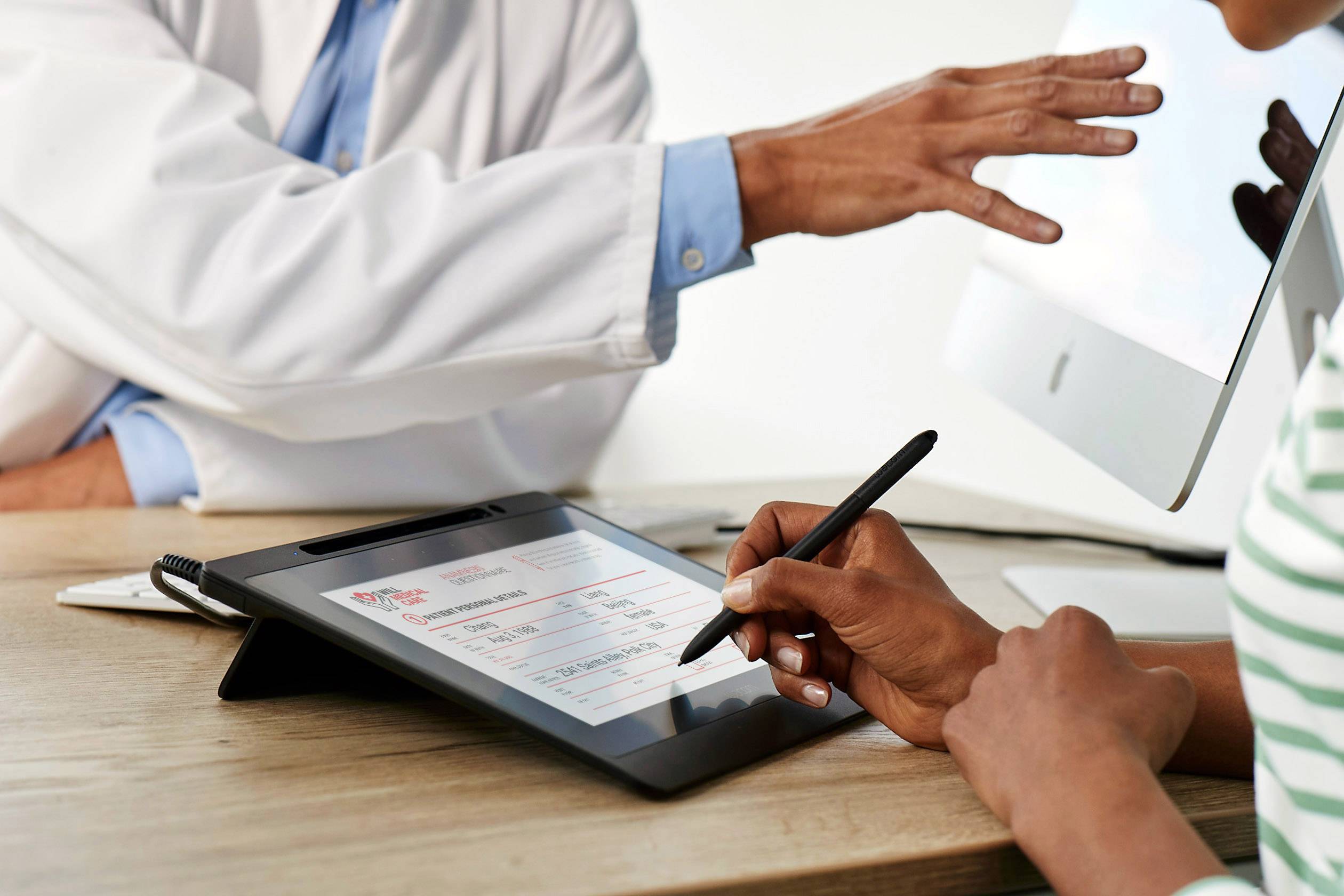 A person is writing on a tablet with a stylus, while another person in a white lab coat points to a computer screen.