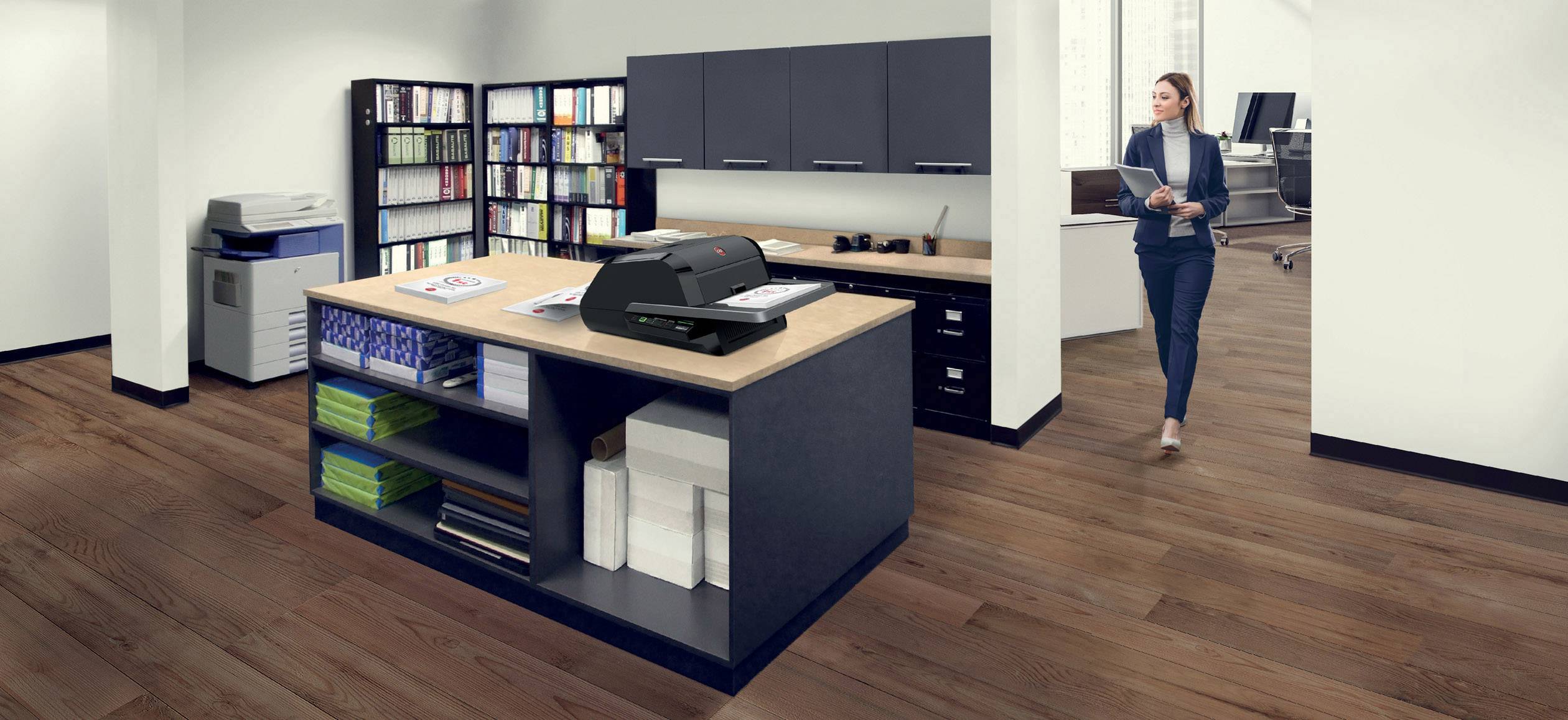 A modern office with wooden flooring, filing cabinets and shelves full of books. One person is carrying documents. A printer sits on a table.