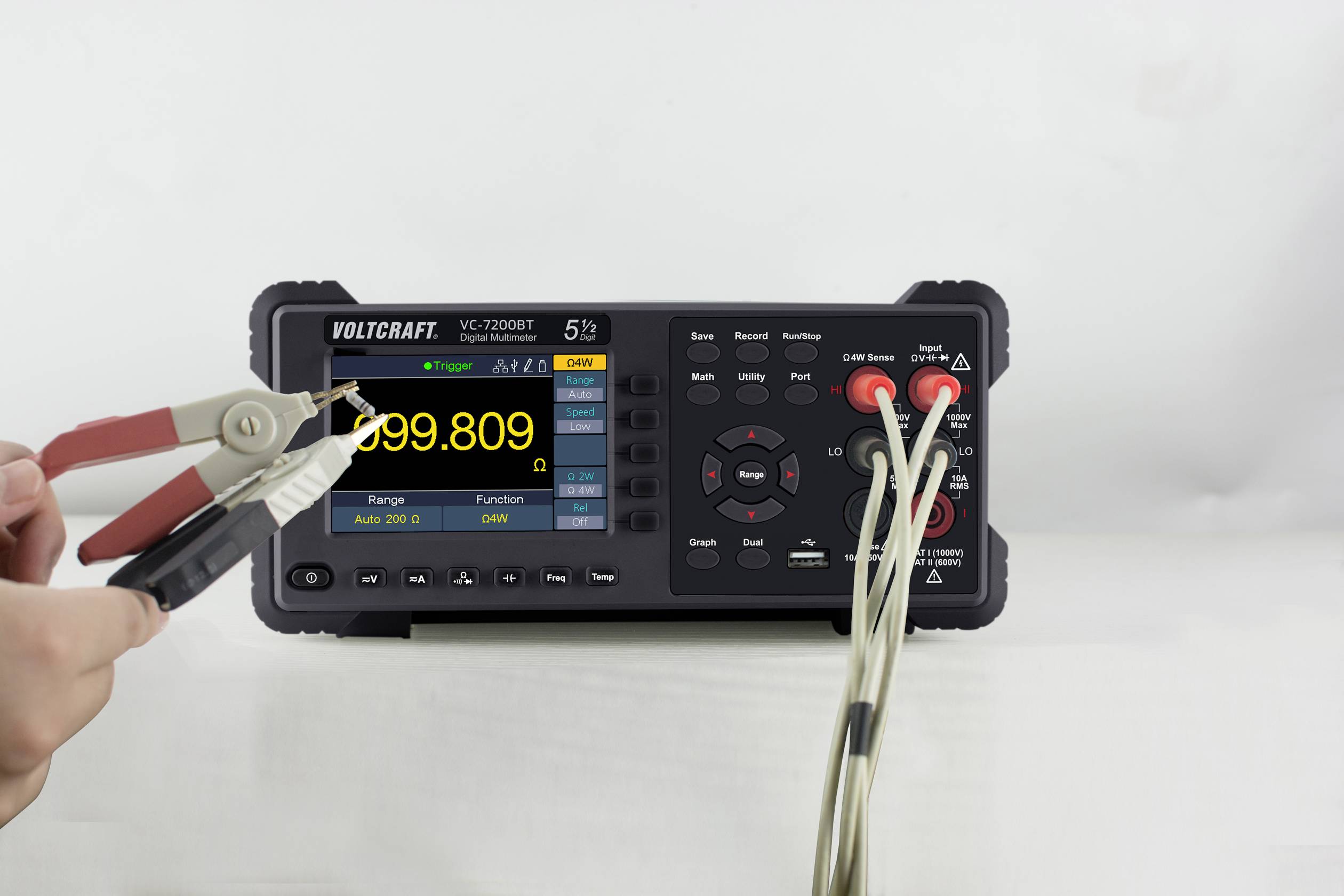A digital multimeter displays a reading of 999.809 on its display. A hand is holding test probes connected by cables.