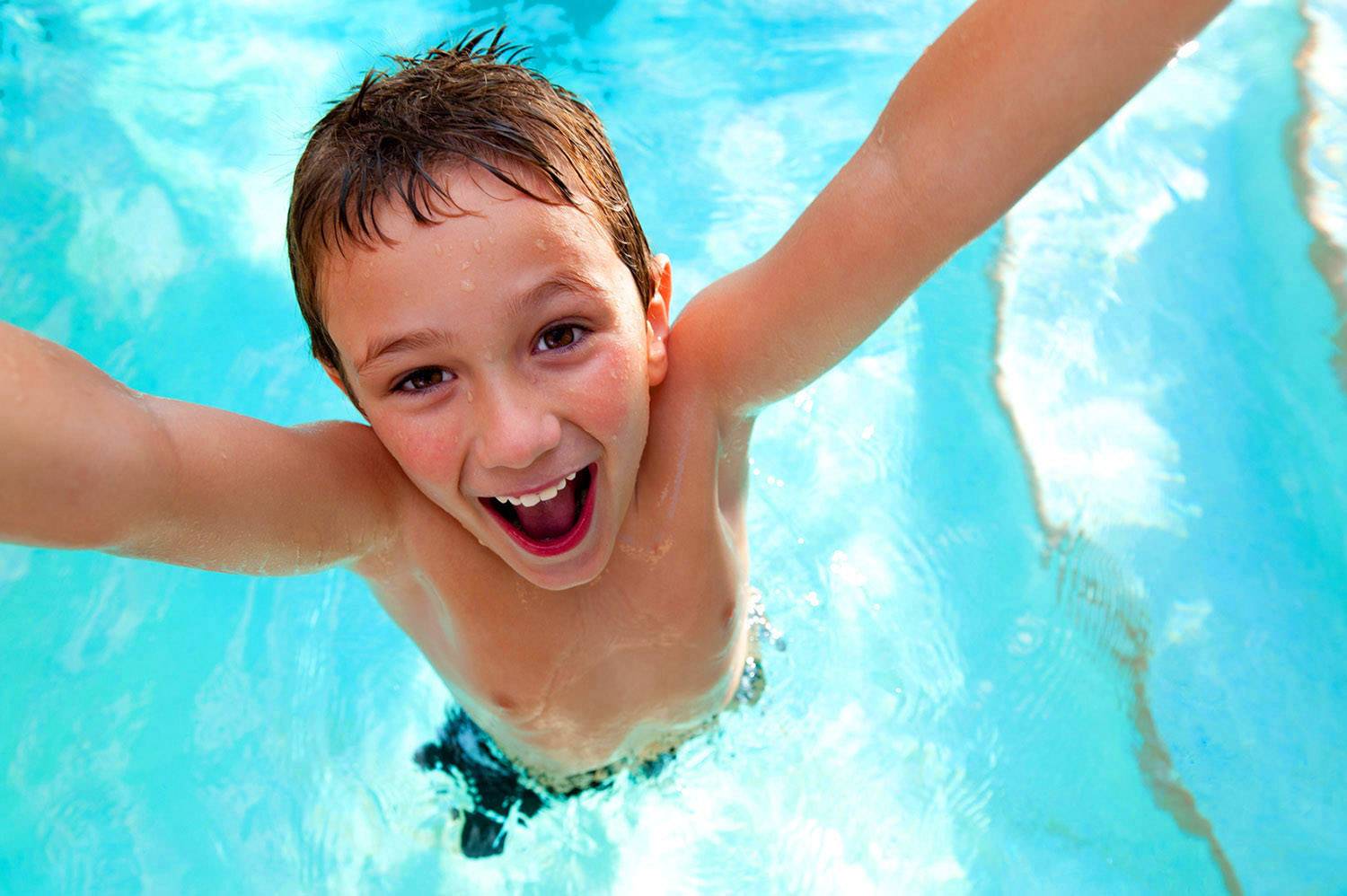 A laughing boy with wet hair swims in the pool and raises his arms joyfully.