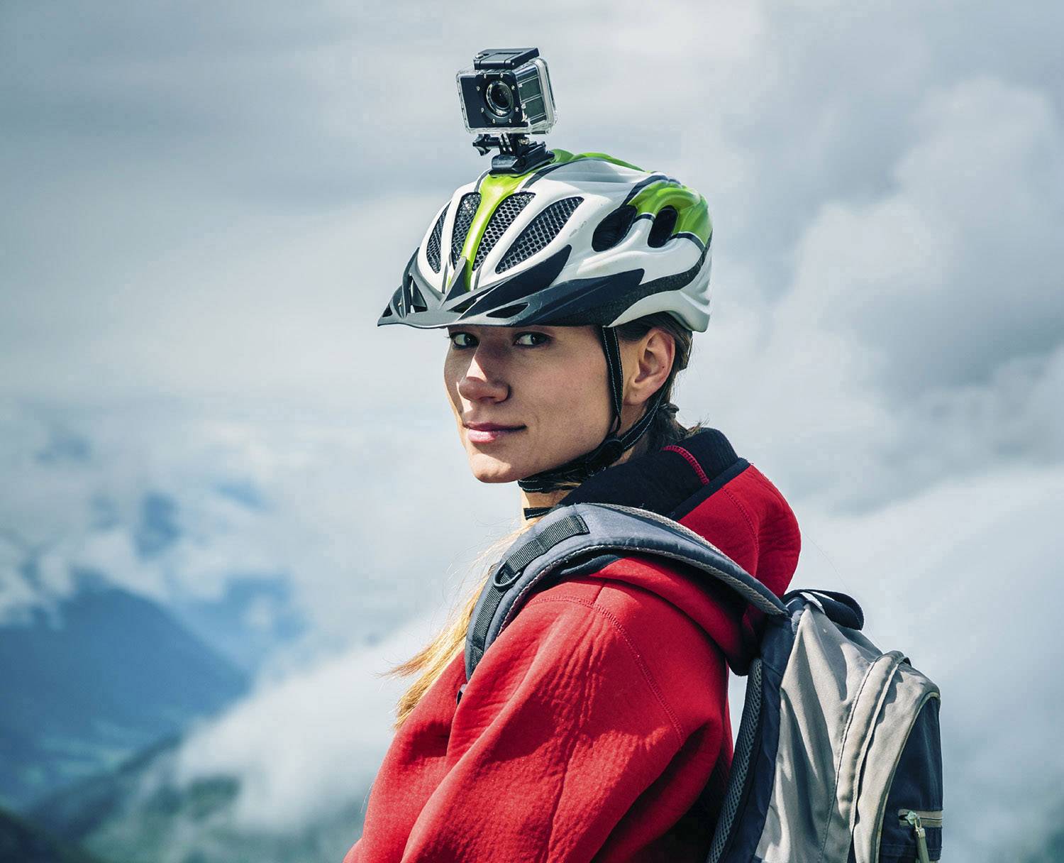 A person wearing a red jumper and helmet, with a camera, stands in front of a mountainous landscape. They are carrying a rucksack and looking to the side.