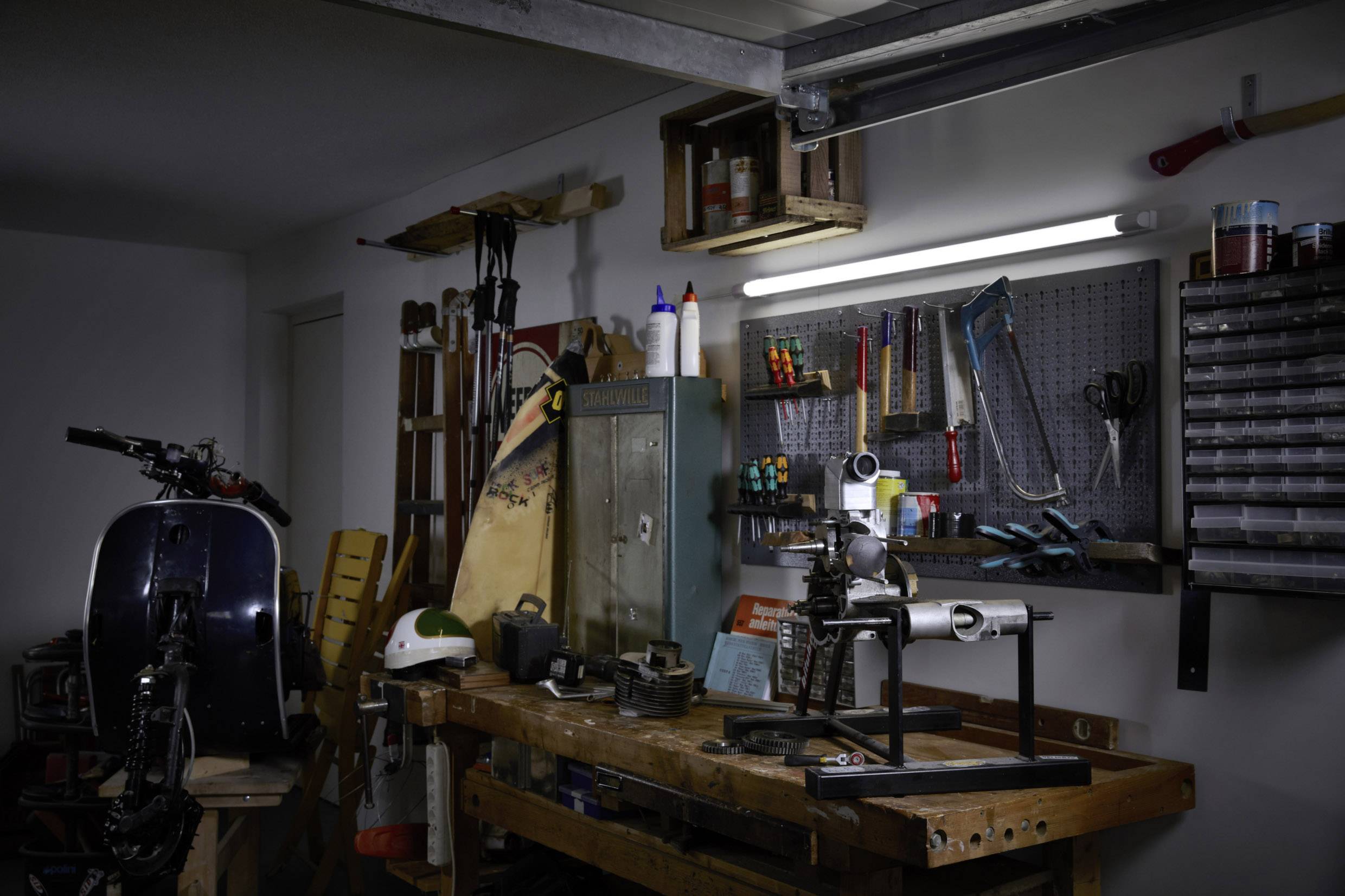Workshop with a workbench, tools on the wall and a moped at the back left. Dim lighting, various craftsman's materials visible.
