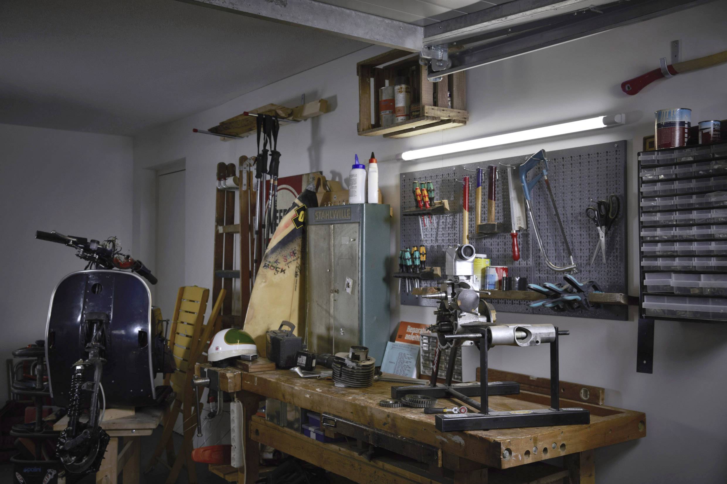 Workshop with a workbench, tools, and materials. In the foreground, a drill; additional tools are hanging on the wall.