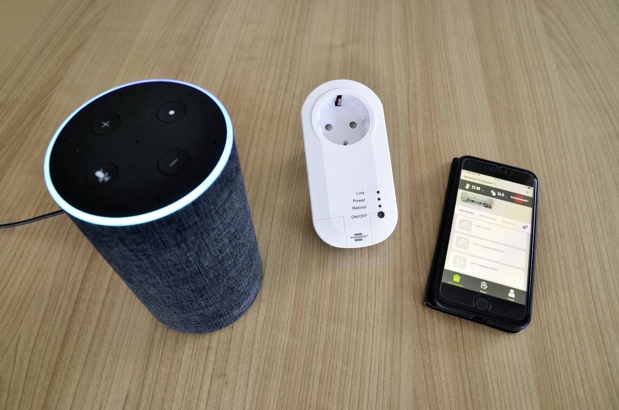 A smart speaker, a smart plug and a smartphone on a table. The smartphone displays a control app.