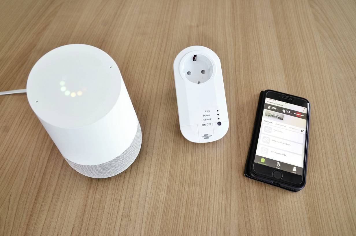 A white smart speaker, a smart plug, and a smartphone on a wooden table. The smartphone displays a smart control app.