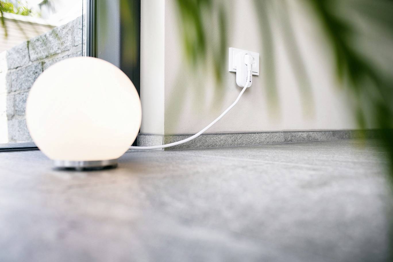 A round, glowing lamp stands on the floor near a window, with a cable plugged into a socket on the wall.