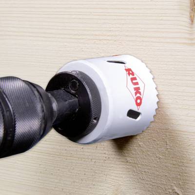 A drill with a white hole saw attachment is cutting into a wooden surface, creating a circular hole.